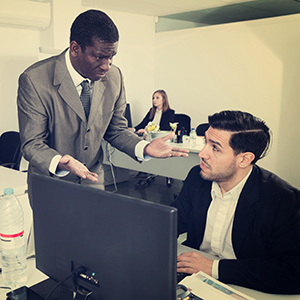 Office scene with one employee gesturing in concern while another listens at a desk computer